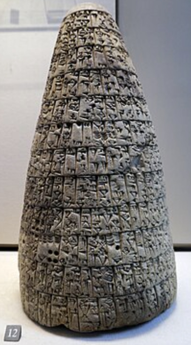 A tall, conical clay artifact densely covered in cuneiform script arranged in horizontal lines, displayed upright in a museum setting with a label "12" at the base.